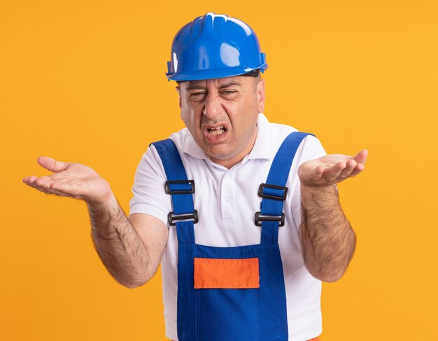 Annoyed adult builder man in uniform holds hands open isolated on orange wall