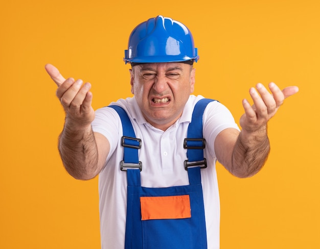 Annoyed adult builder man in uniform points at front with two hands isolated on orange wall