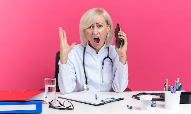 annoyed adult female doctor in medical robe with stethoscope sitting at desk with office tools holding phone and yelling at someone isolated on pink wall with copy space