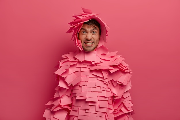 Free photo annoyed adult man clenches teeth, makes angry grimace, covered with sticky notes, has creative costume, clenches teeth, isolated over pink wall, frowns face. human face expressions concept