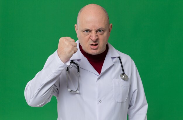 Annoyed adult slavic man in doctor uniform with stethoscope clenching his fist