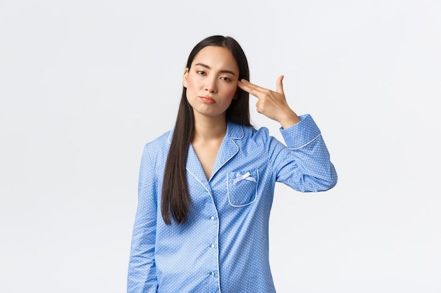 Annoyed and bothered asian girl in blue pajamas looking with reluctant, shooting herself with gun gesture as feeling fed up, tired of hearing or seeing something boring or dumb, white background.