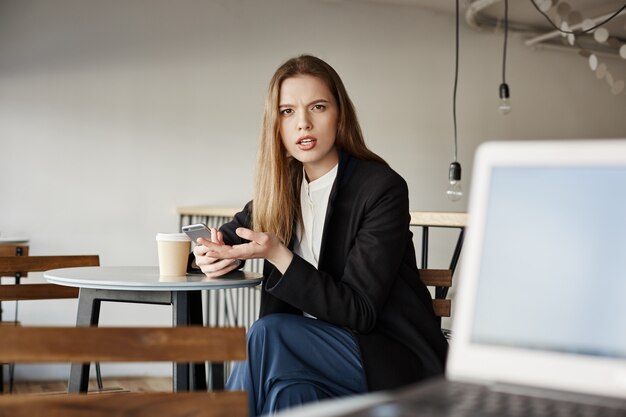 Annoyed businesswoman sit in cafe with mobile phone and looking at person irritated