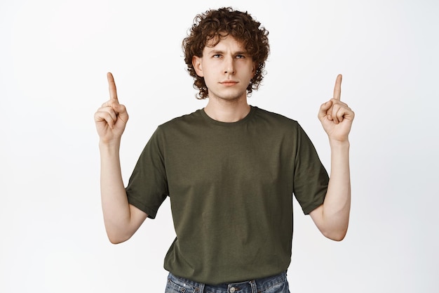 Free photo annoyed curlyhaired man pointing fingers up looking at logo reading advertisement with irritated face expression white background