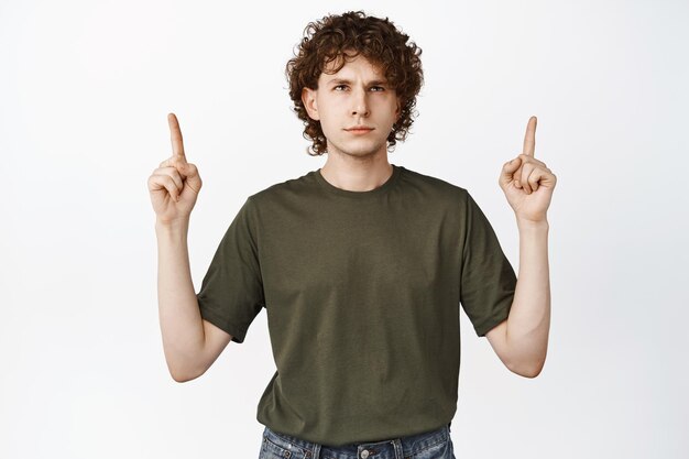 Annoyed curlyhaired man pointing fingers up looking at logo reading advertisement with irritated face expression white background