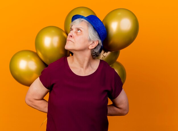 Annoyed elderly woman wearing party hat holds helium balloons behind looking up on orange