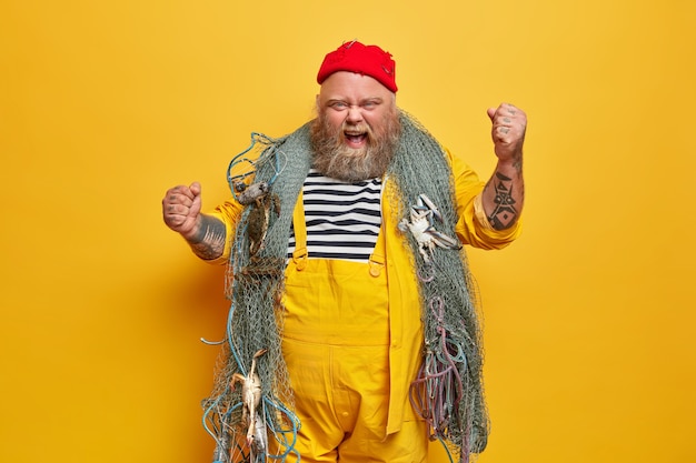 Free Photo annoyed emotional male sailor or professional seaman has sea cruise poses with fishing net raises tattooed arms and yells outraged wears red hat and yellow overalls stands indoor. marine life concept