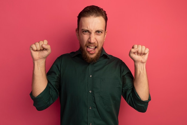 Free photo annoyed handsome blonde man stucks out tongue and stands with raised fists isolated on pink wall