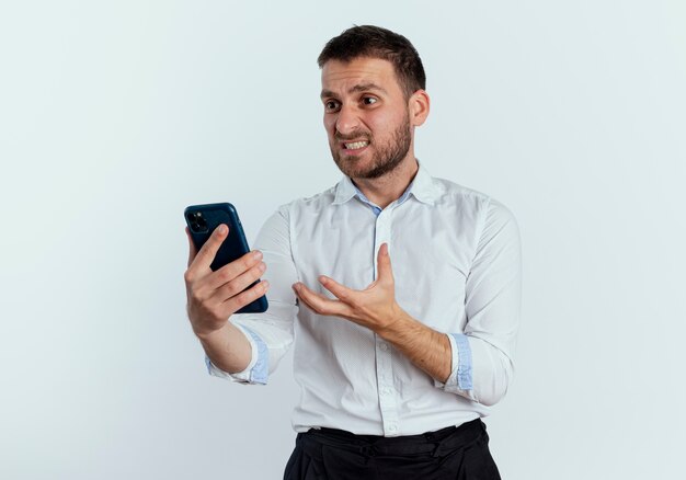 Annoyed handsome man holds and looks at phone raising hand isolated on white wall