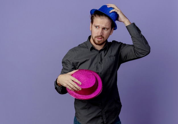 Free photo annoyed handsome man holds pink hat and puts on head blue hat isolated on purple wall