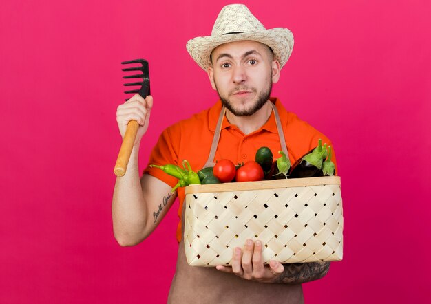 Annoyed male gardener wearing gardening hat holds vegetable basket and rake 