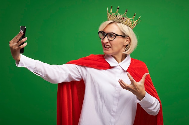 Free photo annoyed middle-aged blonde superhero woman in red cape wearing glasses and crown keeping hand in air taking selfie isolated on green wall