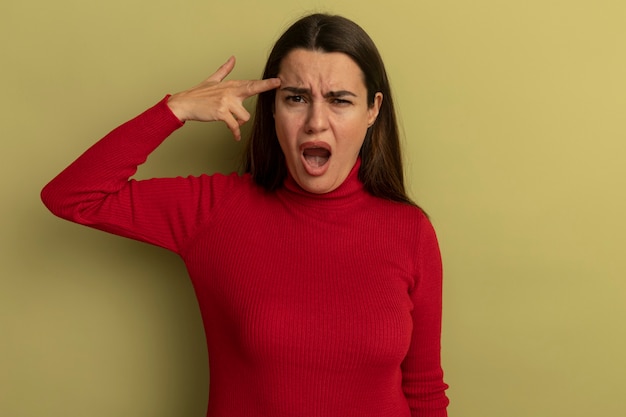 Annoyed pretty caucasian woman gestures gun hand sign putting at temple on olive green