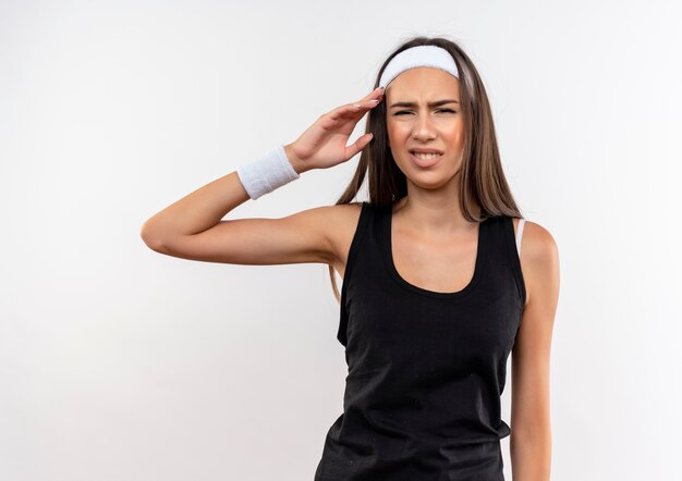 Annoyed pretty sporty girl wearing headband and wristband putting hand near head isolated on white wall