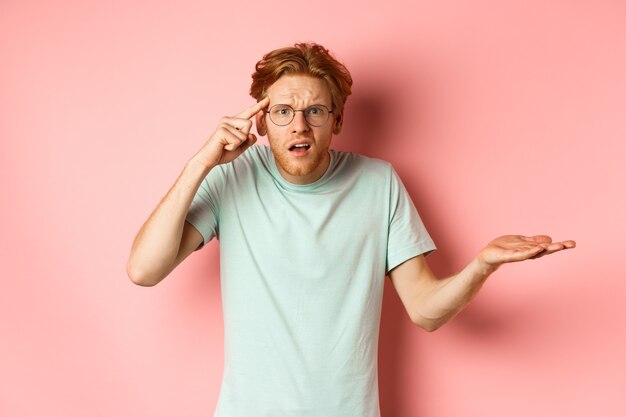 Annoyed redhead man scolding someone stupid pointing finger at head and shrugging standing over pink...