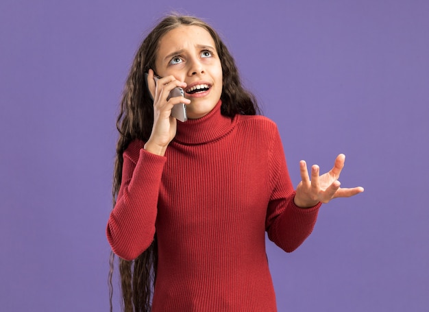 Annoyed teenage girl talking on phone looking up showing empty hand isolated on purple wall