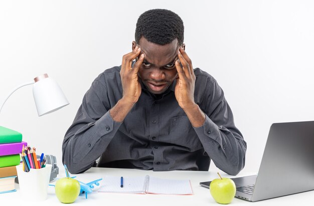 Annoyed young afro-american student sitting at desk with school tools putting hands on his forehead 