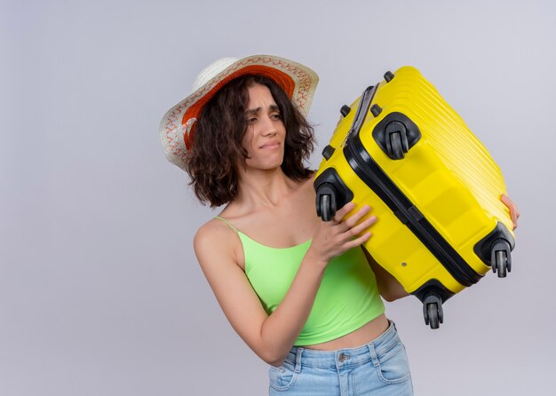 Annoyed young beautiful traveler woman wearing hat and holding suitcase on isolated white wall with copy space