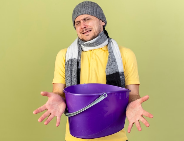 Free Photo annoyed young blonde ill slavic man wearing winter hat and scarf holds plastic bucket isolated on olive green wall with copy space