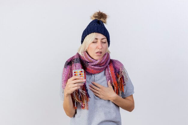 Annoyed young blonde ill woman wearing winter hat and scarf puts hand on chest and holds pack of medical pills isolated on white wall