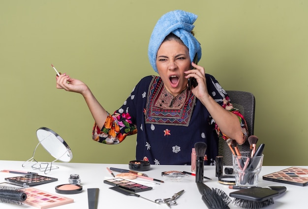 Free Photo annoyed young brunette girl with wrapped hair in towel sitting at table with makeup tools yelling at someone on phone and holding lip gloss isolated on olive green wall with copy space