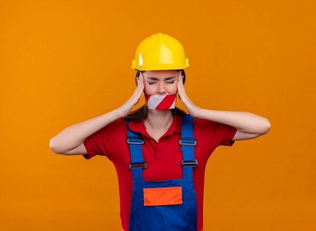 Free photo annoyed young builder girl mouth sealed with warning tape holds head with both hands on isolated orange background