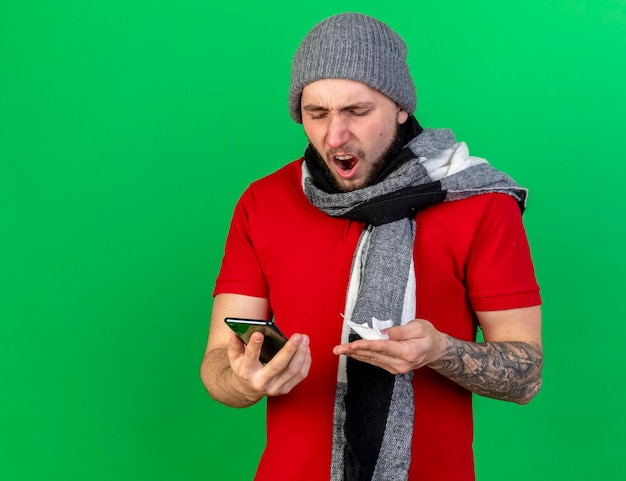 Annoyed young caucasian ill man wearing winter hat and scarf holds tissue