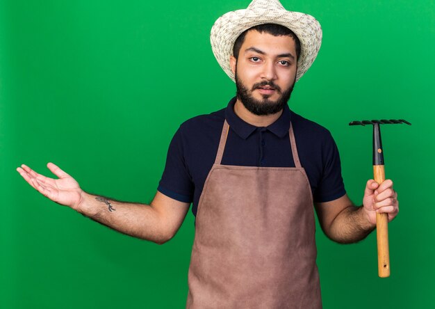 annoyed young caucasian male gardener wearing gardening hat holding rake and keeping hand open isolated on green wall with copy space