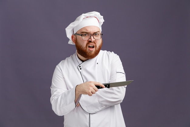 Free photo annoyed young chef wearing glasses uniform and cap looking at camera pointing to side with knife isolated on purple background