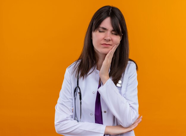 Annoyed young female doctor in medical robe with stethoscope puts hand on cheek on isolated orange background with copy space