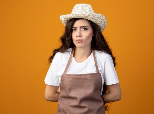 Annoyed young female gardener in uniform wearing gardening hat holds hands behind back isolated on orange wall with copy space