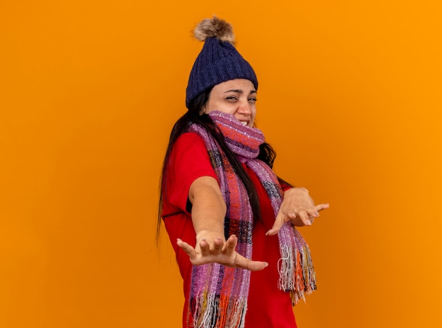 Free Photo annoyed young ill woman wearing winter hat and scarf standing in profile view stretching out hands towards front looking at front gesturing no isolated on orange wall