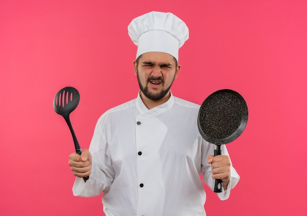 Free photo annoyed young male cook in chef uniform holding frying pan and slotted spoon isolated on pink wall