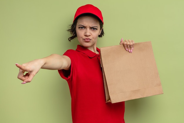 Annoyed young pretty delivery woman holding paper food bag and pointing at side