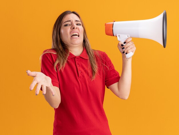 Annoyed young pretty woman holds loud speaker and points at front with hand isolated on orange wall