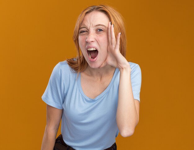 Annoyed young redhead ginger girl with freckles puts hand on face isolated on orange wall with copy space