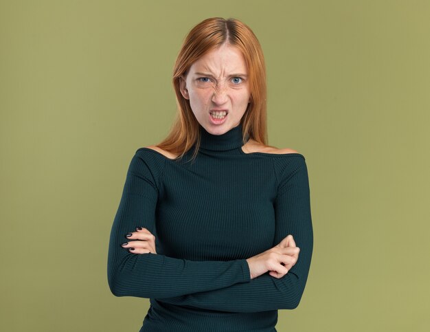 Annoyed young redhead ginger girl with freckles standing with crossed arms isolated on olive green wall with copy space