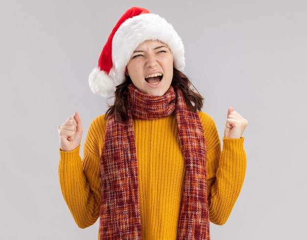 Annoyed young slavic girl with santa hat and with scarf around neck keeps fists and looks at side isolated on white wall with copy space