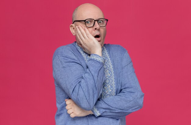 Anxious adult man in blue shirt wearing glasses putting hand on his face and looking 