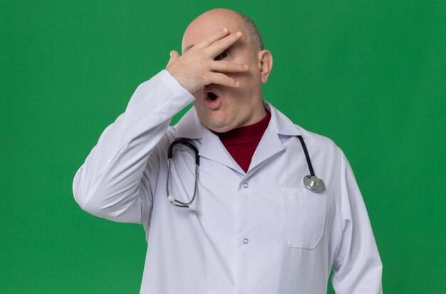 Anxious adult man in doctor uniform with stethoscope putting hand on his face and looking through fingers 