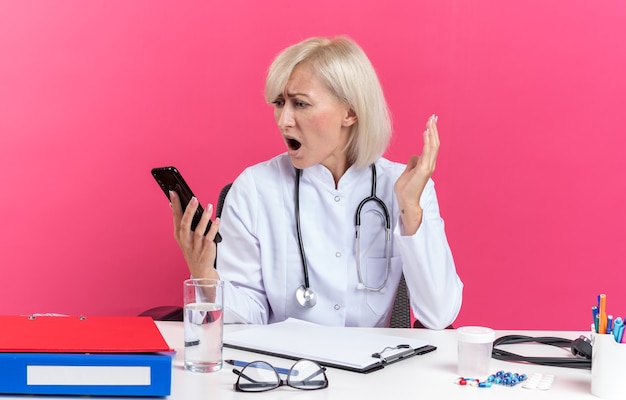 Free Photo anxious adult slavic female doctor in medical robe with stethoscope sitting at desk with office tools holding and looking at phone isolated on pink background with copy space