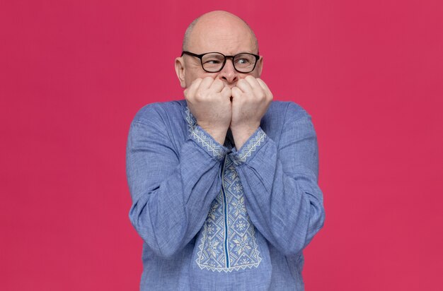 Anxious adult slavic man in blue shirt wearing optical glasses biting his nails