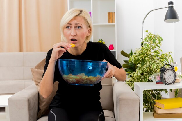 Anxious beautiful blonde woman sits on armchair holding and eating bowl of chips