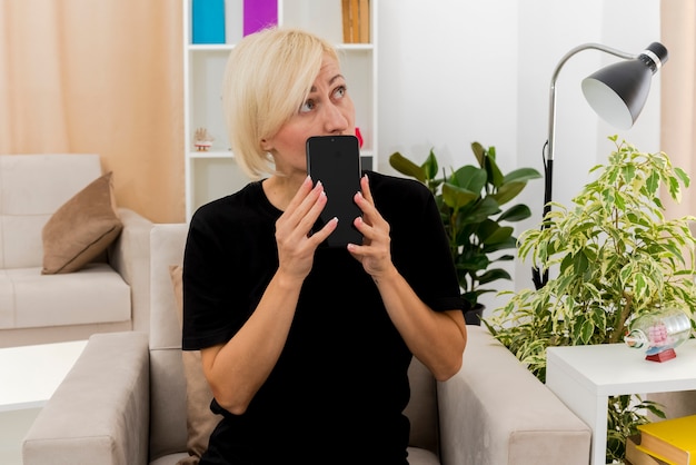 Anxious beautiful blonde woman sits on armchair holding phone and looking at side