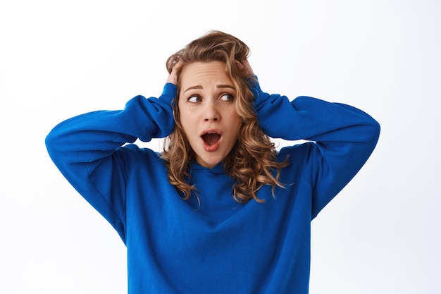 Anxious blond girl panicking, holding hands on head and screaming alarmed, look aside at promotional text with troubled worried face, white wall