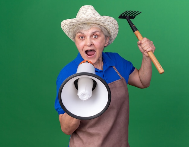 Anxious elderly female gardener wearing gardening hat holding rake and shouting into loud speaker isolated on green wall with copy space