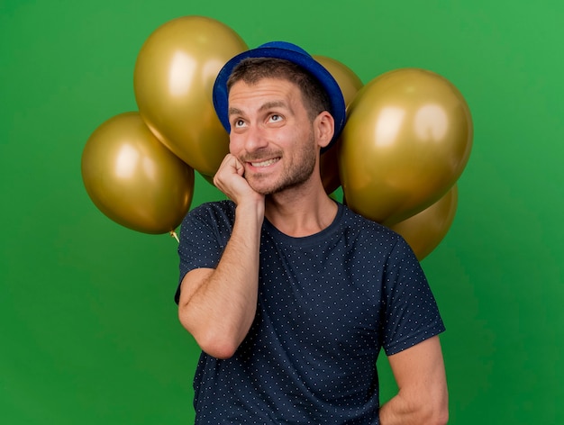 Anxious handsome man wearing blue party hat puts hand on chin looking up holds helium balloons isolated on green wall