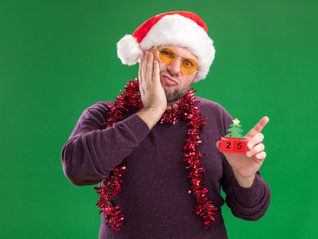 Free Photo anxious middle-aged man wearing santa hat and tinsel garland around neck with glasses holding christmas tree toy with date keeping hand on face looking at camera isolated on green background