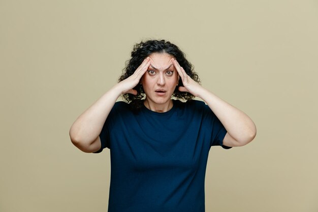 Anxious middleaged woman wearing tshirt keeping hands on head looking at camera isolated on olive green background