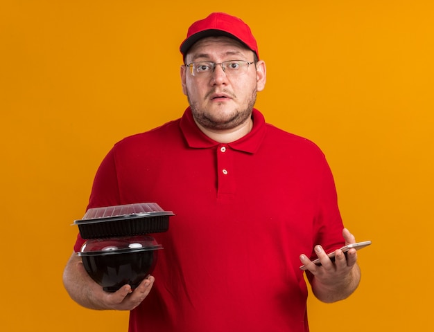 anxious overweight young deliveryman in optical glasses holding food containers and phone isolated on orange wall with copy space